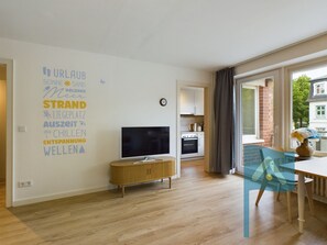 Una sala de estar moderna con un televisor de pantalla plana, piso de madera, un comedor con mesa y sillas, y una cocina visible a través de una puerta abierta.