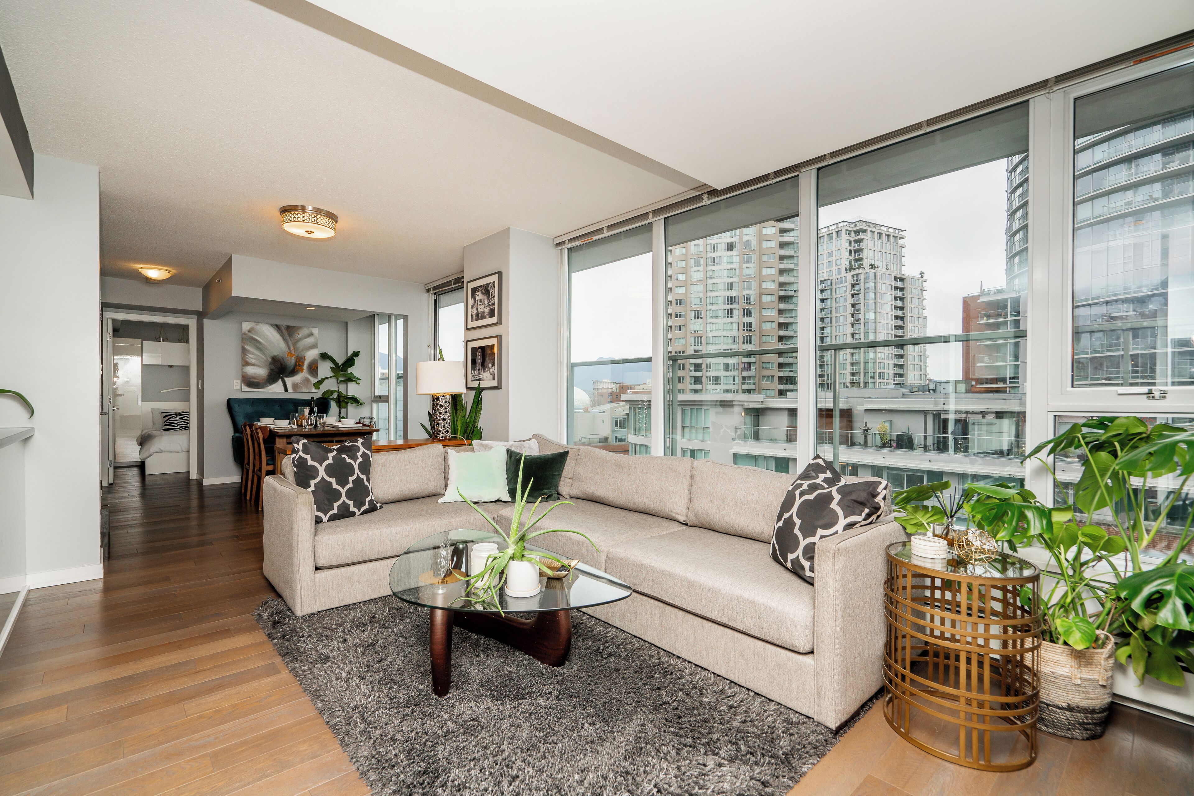 A modern living room with a beige sofa, a glass coffee table, and large windows offering a view of high-rise buildings.