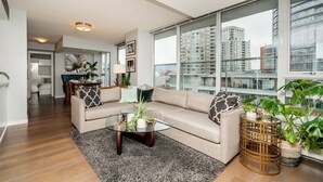 Una sala de estar moderna con un sofá beige, una mesa de centro de vidrio y amplias ventanas con vista a edificios altos.
