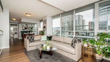 A modern living room with a beige sofa, a glass coffee table, and large windows offering a view of high-rise buildings.