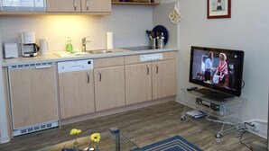 A compact kitchen with wooden cabinets, a microwave, a coffee maker, and a television on a cart. - Haus Teunis - Vacation in the best location, in the pedestrian zone in Wyk - Teunis (Unknown)