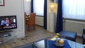A hotel room with a television, a dining table with chairs, a small cabinet, a radiator, and a window with curtains. - Haus Teunis - Vacation in the best location, in the pedestrian zone in Wyk - Teunis (Unknown)