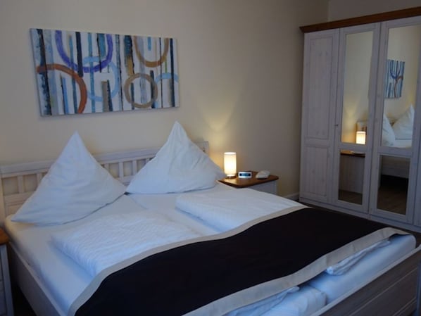 Un lit bien fait, avec du linge de lit blanc et une couverture noire, une table de chevet avec une lampe, et une armoire avec un miroir.