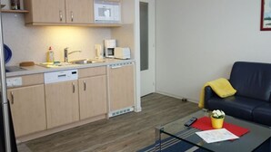 A compact kitchen with wooden cabinets, a microwave, and a coffee maker. There's a glass table with a remote control and a red placemat. A dark blue sofa with a yellow throw pillow is against the wall.