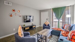 A living room with a grey sofa, a blue sofa, a wooden coffee table, and a TV on the wall. There are decorative plates on the wall and a green curtain over the window.