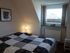 A bedroom with a checkered bedspread, a bedside lamp, a window with curtains, and a radiator.