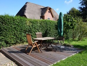 Una terraza de madera con una mesa y sillas, un parasol verde y una casa con techo de paja al fondo.
