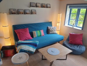 Quarto aconchegante com sofá azul, pequena mesa de madeira, um bean bag e janela com vista para vegetação.