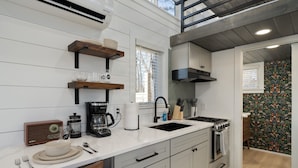 A modern kitchen with white cabinetry, a black stove, and a coffee maker. There are wooden shelves above the sink and a window with blinds.