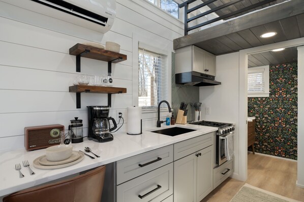 Eine moderne Küche mit weißen Schränken, einem schwarzen Herd und einer Kaffeemaschine. Darüber befinden sich Holzregale über der Spüle, und ein Fenster mit Jalousien ist zu sehen.