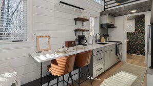 A modern kitchen with white cabinetry, a white countertop, and a built-in stove. There are two bar stools, a coffee maker, and a framed picture on the counter.
