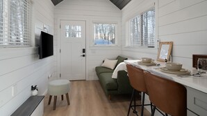 A compact kitchen with white paneling, a wooden ceiling, a green sofa, a bar stool, and a dining area with a table set for two.