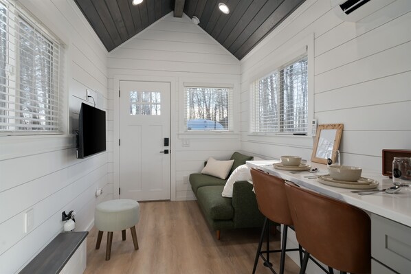 Une cuisine compacte avec un lambris blanc, un plafond en bois, un canapé vert, un tabouret de bar et un coin repas avec une table dressée pour deux personnes.
