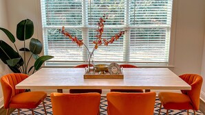 Een eetgedeelte met een houten tafel, oranje stoelen, een kamerbreed tapijt en een groot raam met jaloezieën.