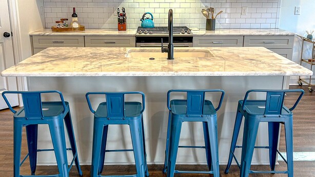 Een keuken met een centraal eiland, vier blauwe barkrukken, witte kasten, een roestvrijstalen afzuigkap en een betegelde achterwand.