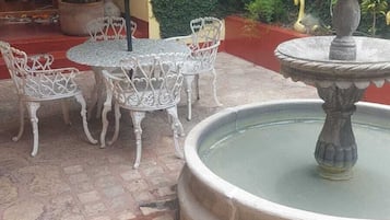 Un patio exterior con una fuente, sillas de metal blanco y una sombrilla verde.
