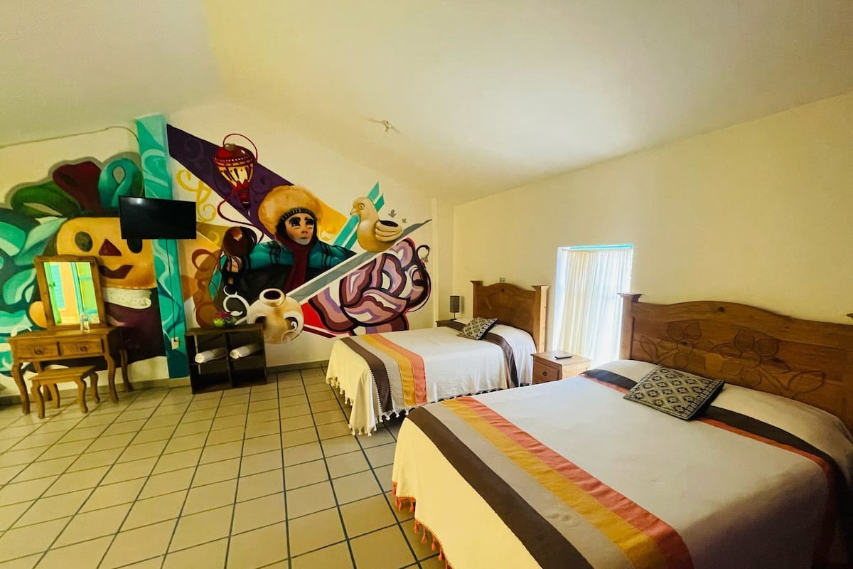A hotel room with two beds, a desk, a chair, a mirror, and a mural on the wall.