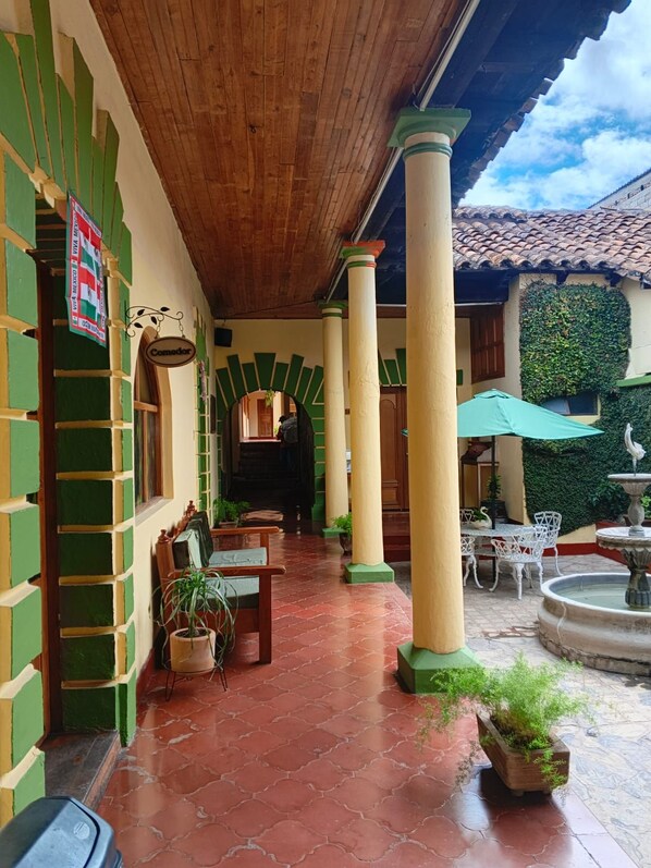 Un espacio al aire libre techado, con piso de baldosas, columnas de madera y una fuente.