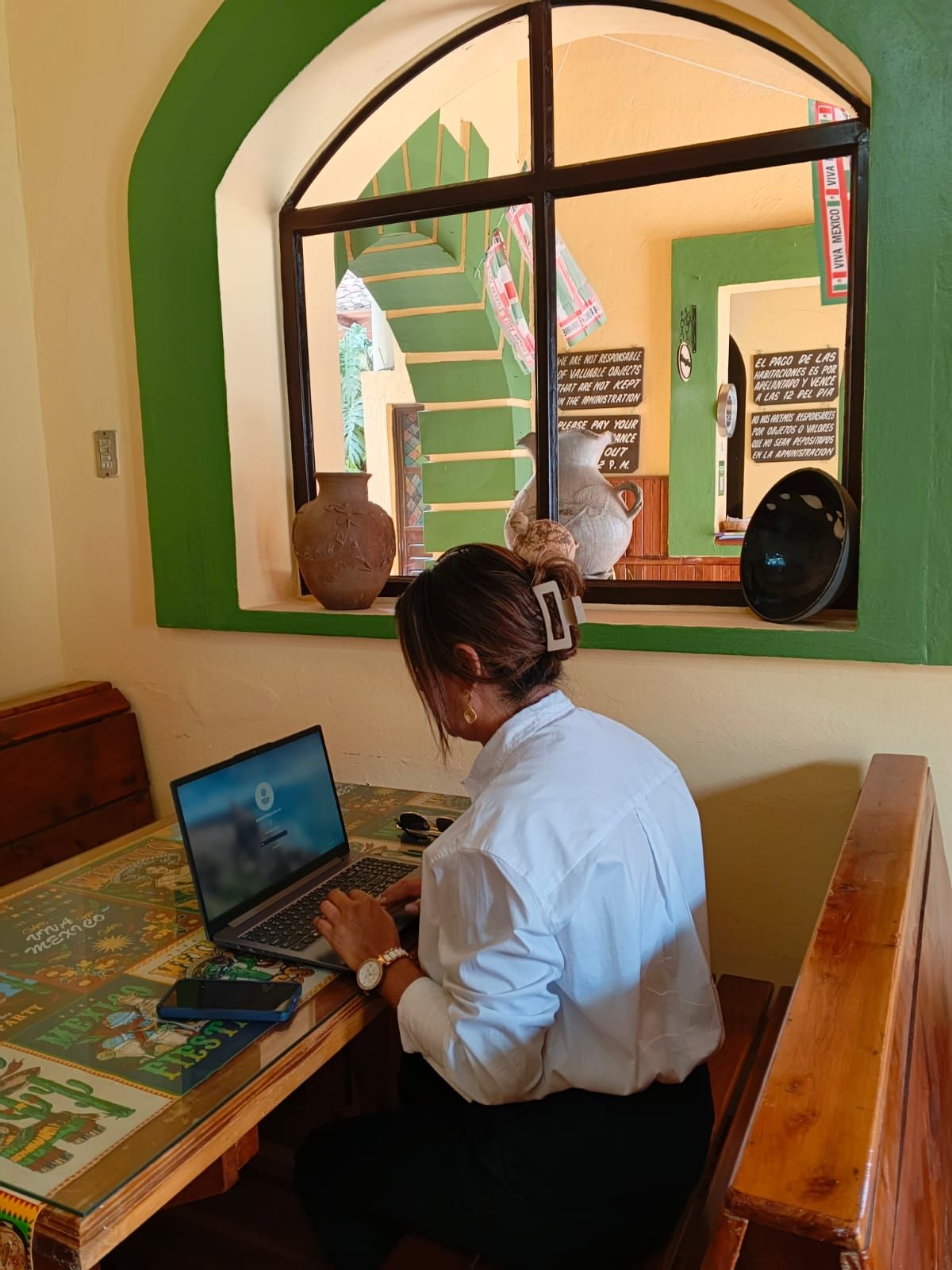 Una persona trabajando en un escritorio de madera con una computadora portátil, en una habitación con un arco ventana verde y vistas a una escalera.
