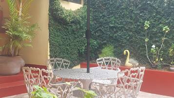 Un patio al aire libre con una mesa redonda, sillas blancas, un paraguas verde y plantas en macetas.