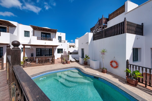 Zona de piscina con terraza de madera, edificios blancos y un flotador en la pared.