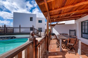 Una terraza de madera con piscina, mobiliario exterior y vistas a edificios blancos.