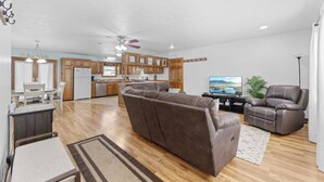 A spacious living area with a brown leather sofa, a dining table with chairs, a kitchen in the background, and a ceiling fan.