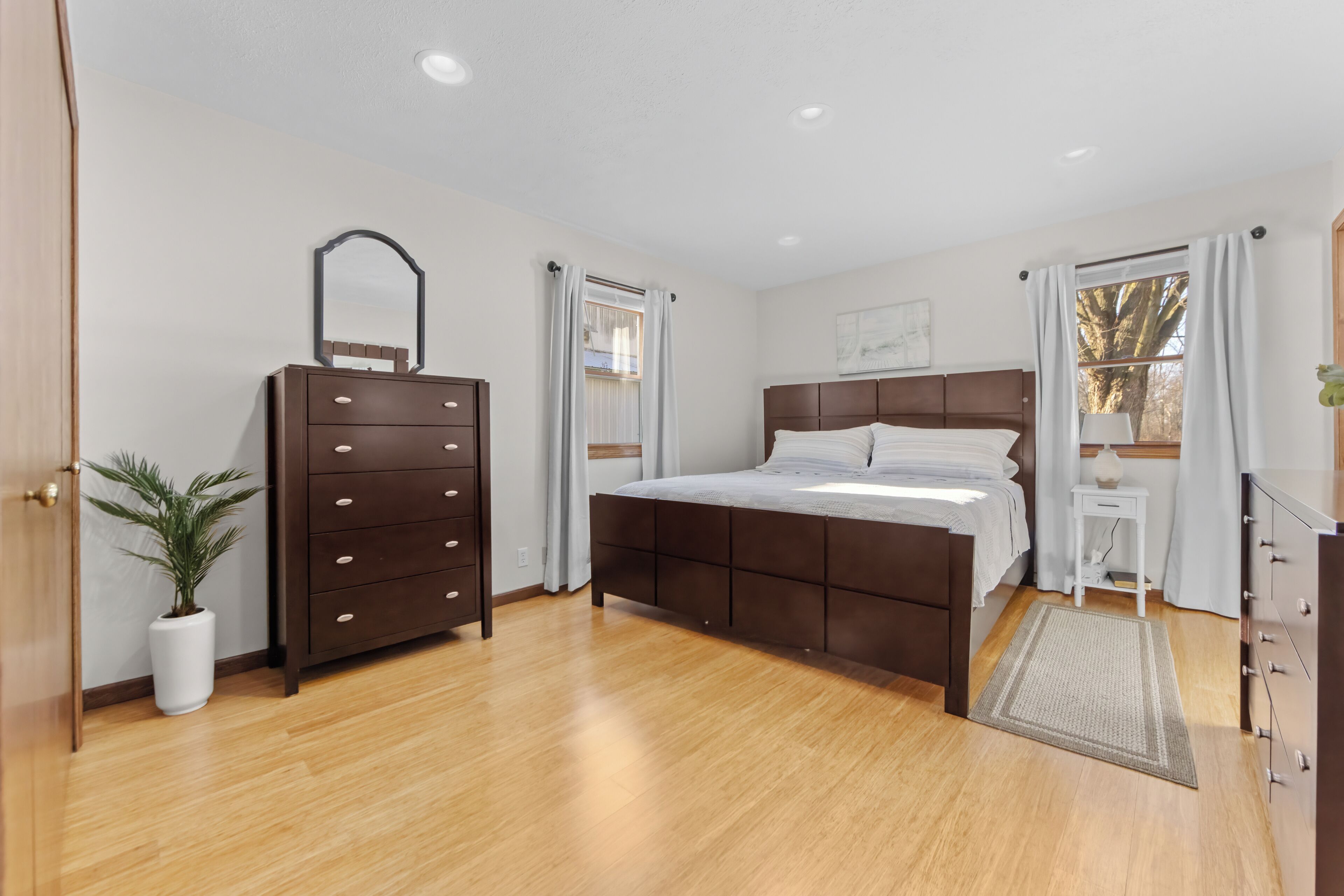 Een slaapkamer met een houten vloer, een bed met hoofdeinde, een dressoir met lades, een spiegel, een tafeltje met een lamp en een potplant.