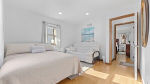A bedroom with a large bed, a smaller sofa, a window with curtains, a painting on the wall, and a door leading to another room.