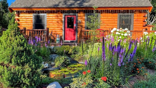 Una cabaña de troncos con puerta roja, rodeada de un jardín con diversas plantas y flores.