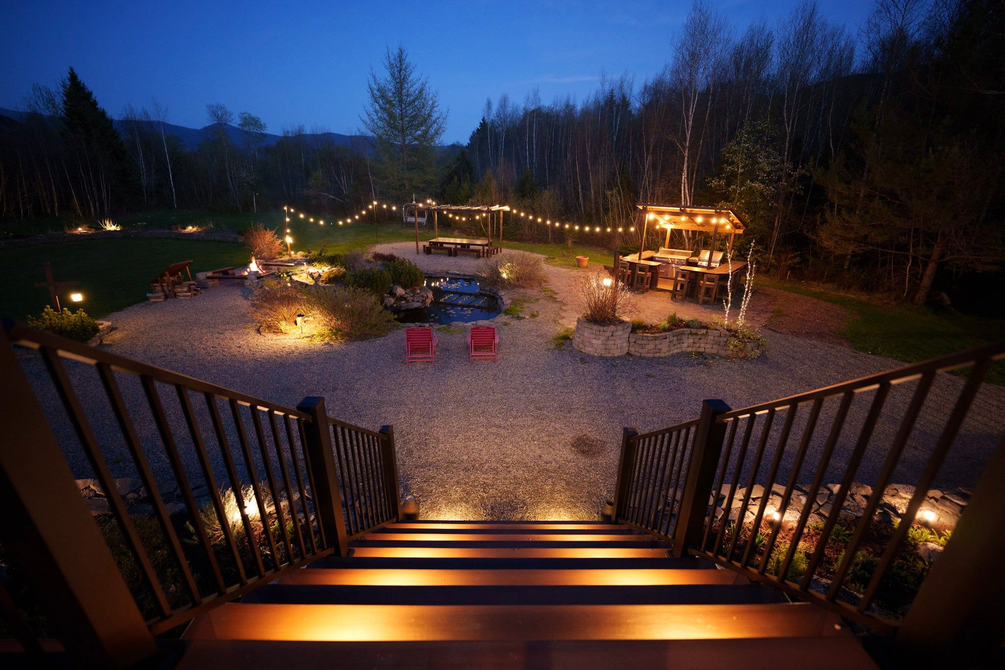 Eine Holztreppe führt zu einer Terrasse mit Außenmöbeln und Grillplatz, umgeben von einem Kiesweg und Bäumen.