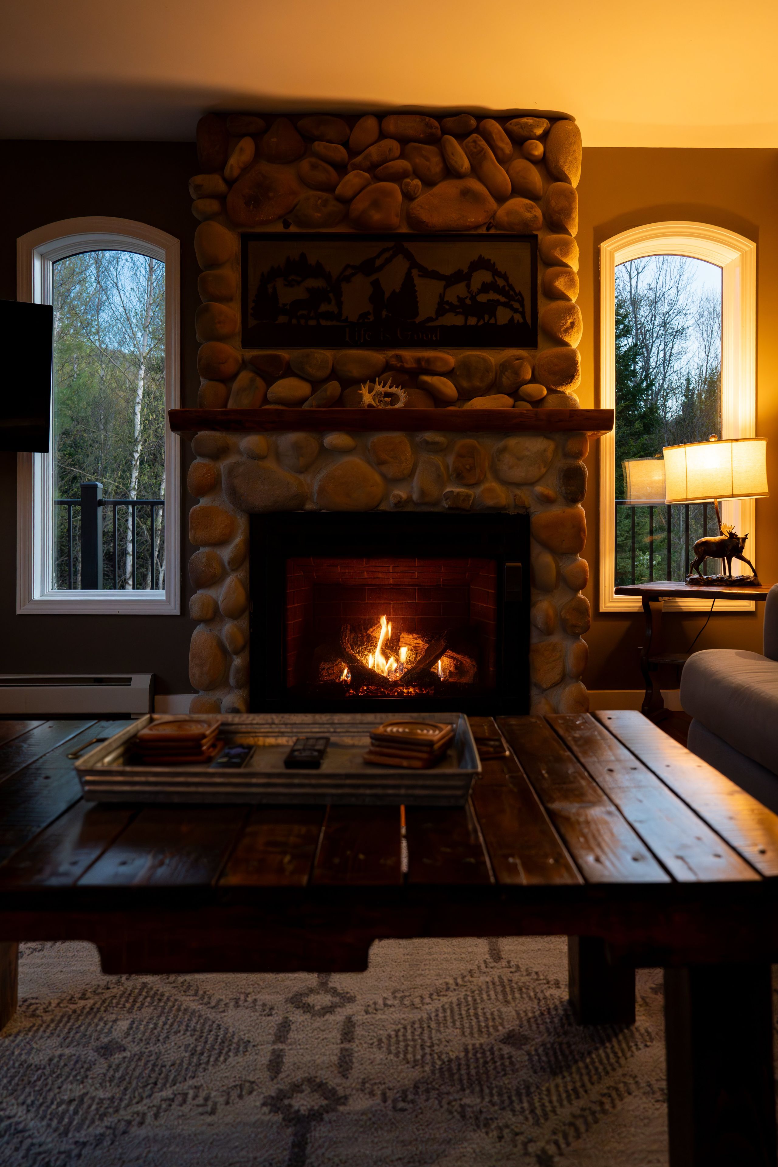 Una chimenea de piedra con fuego, una mesa de centro de madera con libros y mandos a distancia, y una lámpara en una mesita auxiliar.