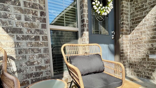 A wicker chair with a cushion and a small table on a porch with a brick wall and a door with a wreath. - Short term rental. Home away from your home. (Celina)
