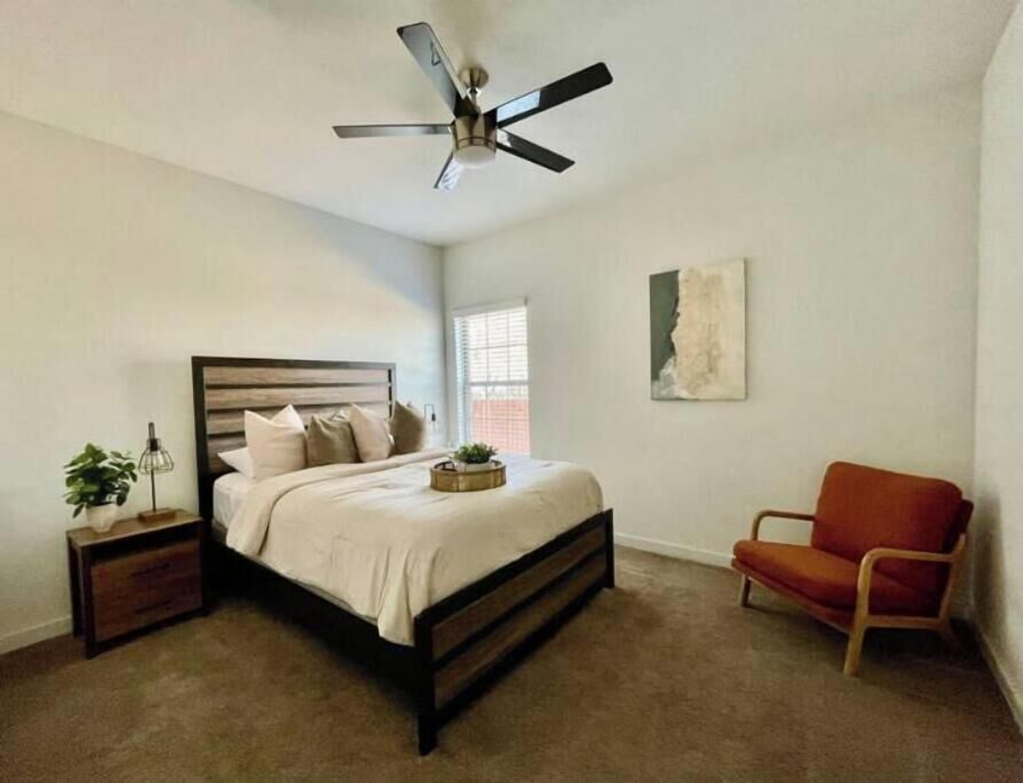 Een slaapkamer met een bed, een stoel, een nachtkastje, een plafondventilator en een raam.
