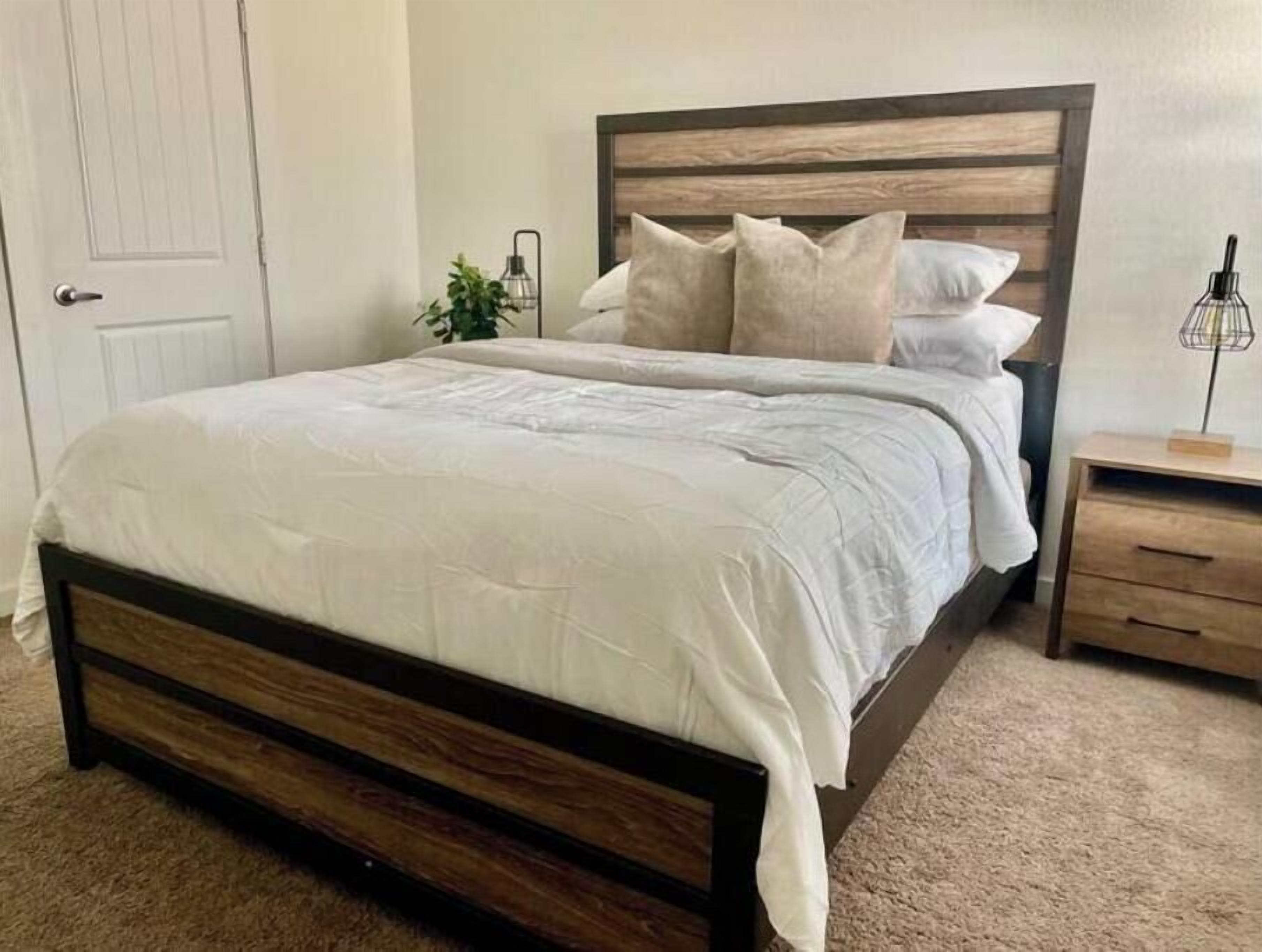 A bedroom with a wooden bed frame, white bedding, a bedside table with a lamp, and a plant.