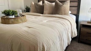 A neatly made bed with a beige comforter and pillows, a woven basket with a plant, and a wooden headboard. - Short term rental. Home away from your home. (Celina)