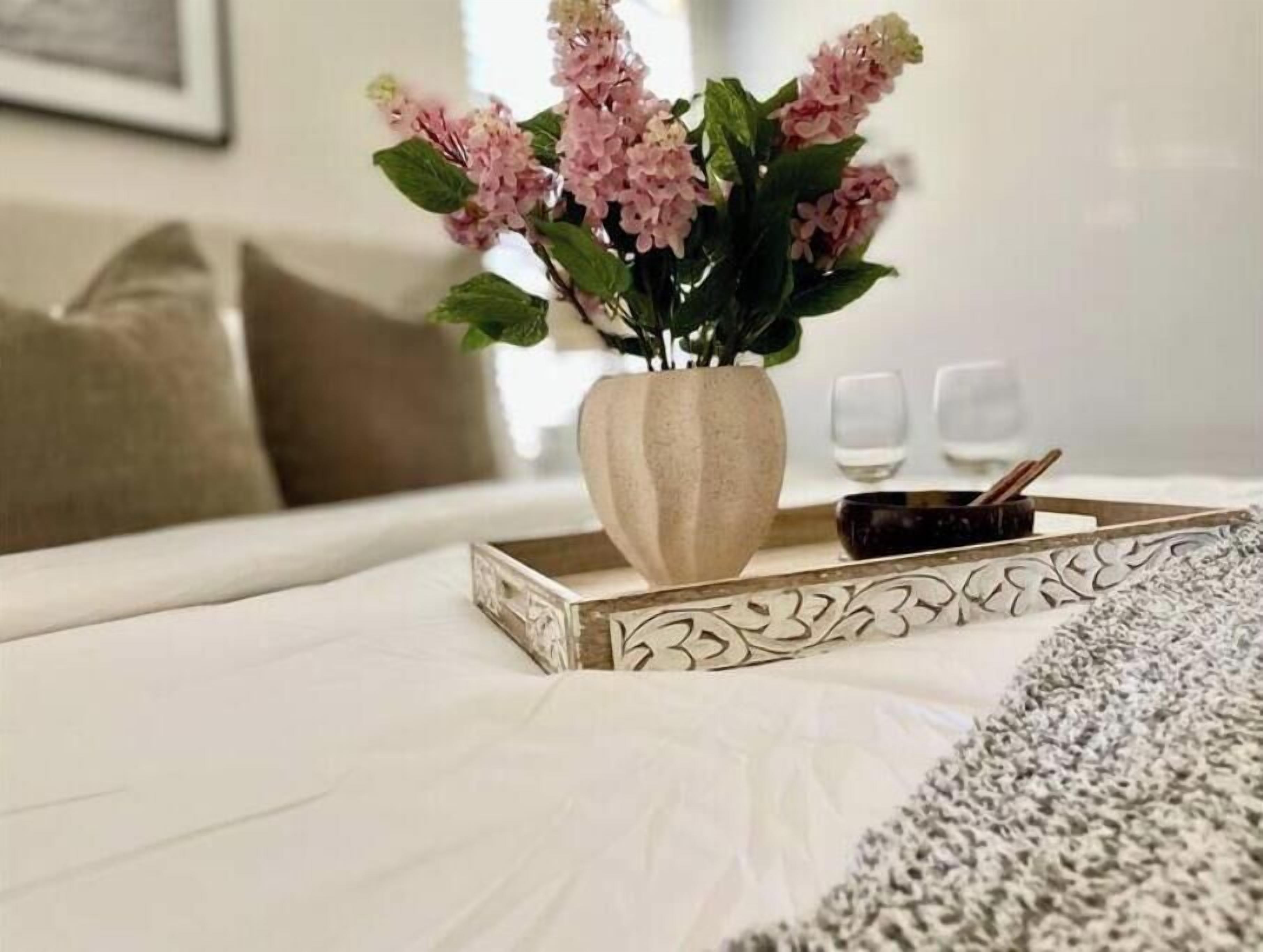 Ein Bett mit weißer Bettdecke, eine Vase mit rosa Blumen, ein Tablett mit einer Kerze und zwei Gläser auf einem Tisch.