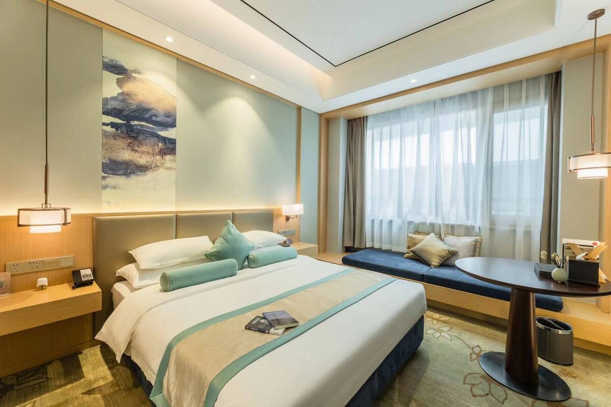 Una habitación de hotel con una cama grande, mesitas de noche, un escritorio y una ventana con cortinas translúcidas.
