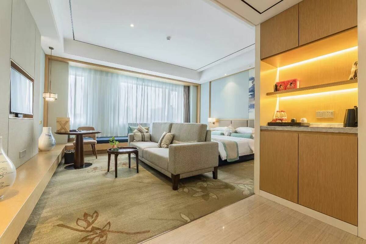 Una habitación de hotel moderna con sofá, una mesita, una mesita de noche, lámpara, ventana con cortinas y un estante empotrado con objetos.