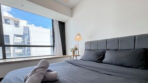 Ein modernes Hotelzimmer mit einem großen Bett, einem Nachttisch mit Lampe und Blick auf Gebäude durch das Fenster.