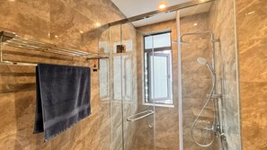 A modern bathroom with a glass shower enclosure, a towel rack with a dark towel, and a window providing natural light. - R&F PRINCESS COVE BY RONGYU (Johor Bahru)