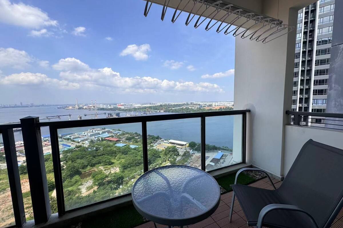A balcony with a glass table and chairs, overlooking a body of water and industrial area.