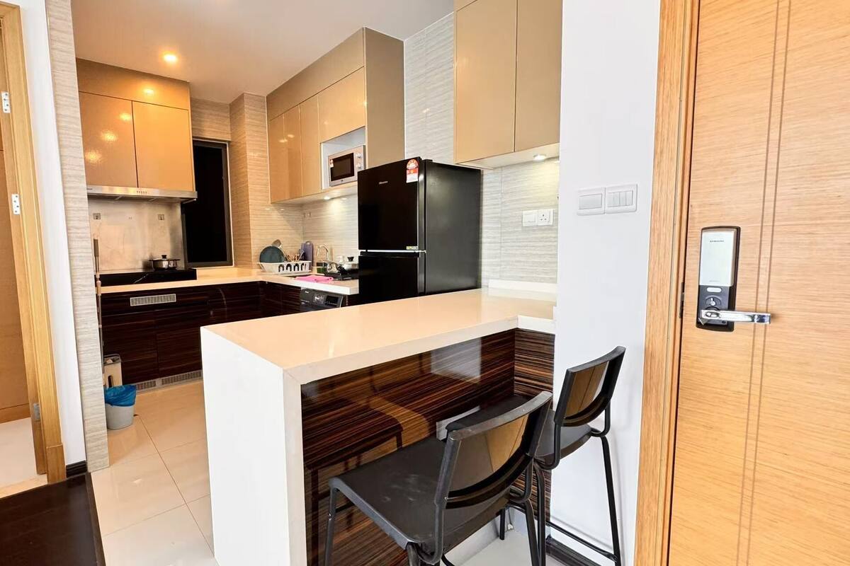 A modern kitchen with dark wood cabinets, a white countertop, and a built-in microwave. There is a black refrigerator and a Samsung smart lock on the door.