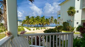 A balcony with a view of a beach, palm trees, and a building with air conditioning units. - Sea View Cabana@SapphireBeachResort (Not available)