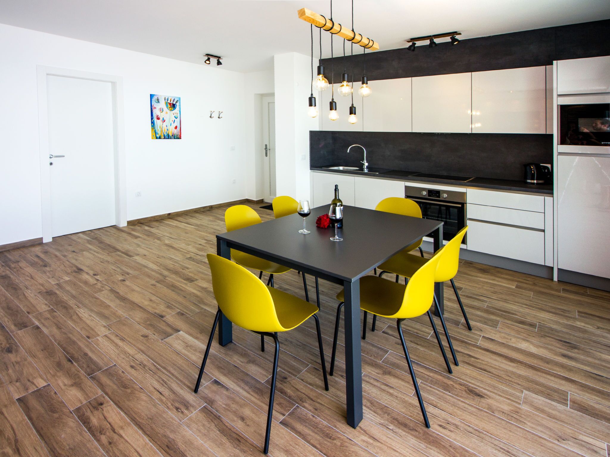 Una cocina moderna con encimera negra, armarios blancos y una zona de comedor con mesa oscura y sillas amarillas.