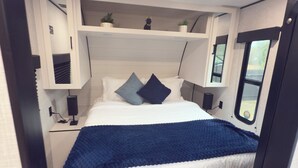A compact bedroom with a bed, bedside tables, a shelf with decorative items, and a window. - Cozy RV Retreat with Private Hot Tub and Fenced Yard (Spring Hill)