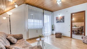 Een gezellige woonkamer met een houten plafond, een bank, een salontafel en een zichtbare eethoek door een open deur.