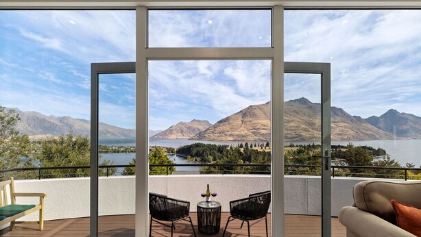 Een balkon met uitzicht op bergen en een meer. Er staan twee stoelen en een tafel met een fles erop.