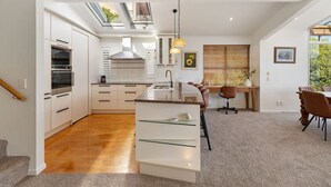 Een moderne keuken met een centraal eiland, een eethoek met een houten tafel en stoelen, en uitzicht op groen via grote ramen.