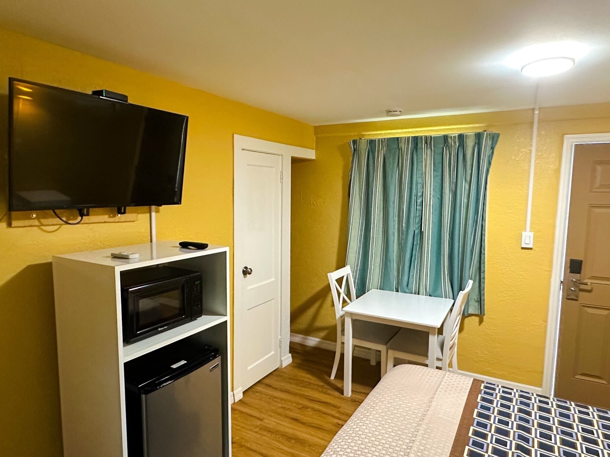 A compact room with a bed, a small kitchenette, a TV mounted on the wall, a door, and a window with curtains.